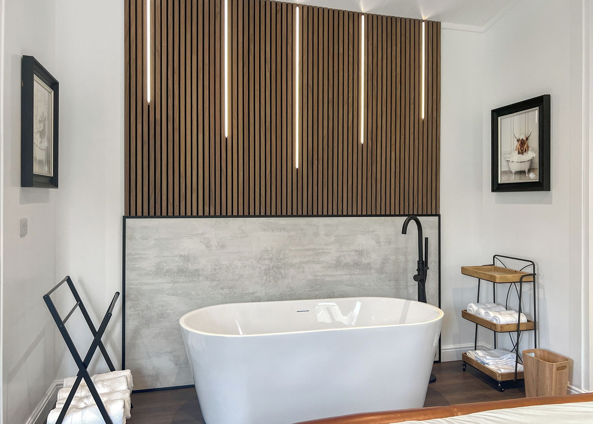 Ein modernes Badezimmer mit einer freistehenden Badewanne, Holzleisten darüber und einem Handtuchhalter mit gefalteten Handtüchern.