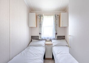 A compact bedroom with two single beds, a small window with curtains, and a wooden headboard.