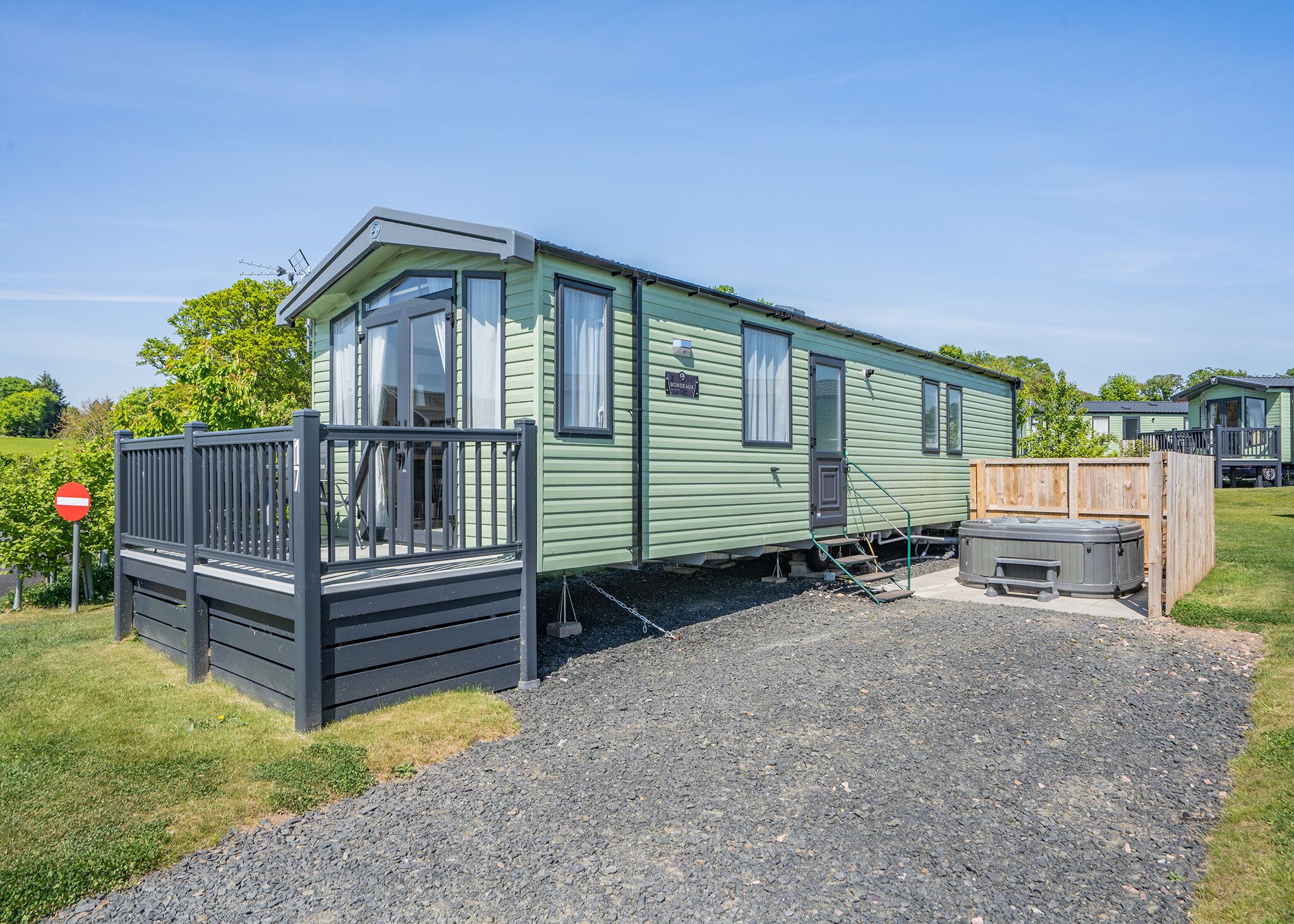 A green mobile home with a fenced porch, a hot tub, and a gravel driveway.