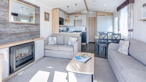 A modern living room with a fireplace, sofa, and coffee table, connected to a kitchen and dining area with wooden accents and a large mirror. - Coldstream Retreat - 3 Bedroom (Pet Friendly) (Coldstream)