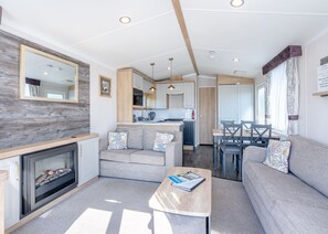 A modern living room with a fireplace, sofa, and coffee table, connected to a kitchen and dining area with wooden accents and a large mirror. - Coldstream Retreat - 3 Bedroom (Pet Friendly) (Coldstream)