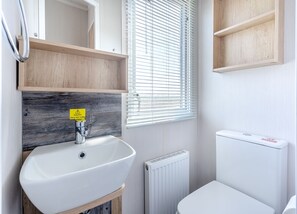 A bathroom with a white sink, wooden shelves, a caution sign, a window with blinds, and a white toilet. - Coldstream Retreat - 3 Bedroom (Pet Friendly) (Coldstream)