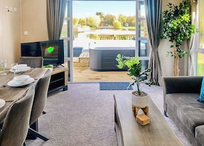 A living room with a TV, dining table, chairs, a sofa, a potted plant, and a view of a hot tub and outdoor area. - Whimbrel Lakeside Lodge (Laceby)