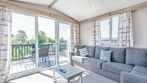A modern living room with a grey sofa, a small table, and a sliding glass door leading to an outdoor deck. - Tweed Retreat - 2 Bedroom (Pet Friendly) (Coldstream)