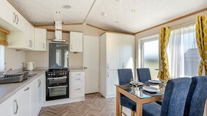 A compact kitchen with modern appliances, a dining area with blue chairs, and a large window with yellow curtains. - Cheviot Retreat - 2 Bedroom (Pet Friendly) (Coldstream)