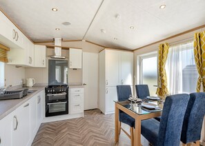 A compact kitchen with modern appliances, a dining area with blue chairs, and a large window with yellow curtains. - Cheviot Retreat - 2 Bedroom (Pet Friendly) (Coldstream)