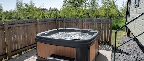 A hot tub with a wooden exterior and a step for access, surrounded by a wooden fence and a house in the background.