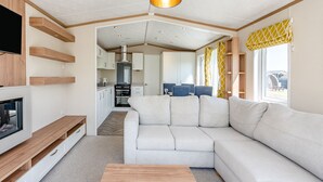 A modern living room with a sectional sofa, a TV, wooden shelves, and a kitchen in the background. - Cheviot Retreat - 2 Bedroom (Pet Friendly) (Coldstream)