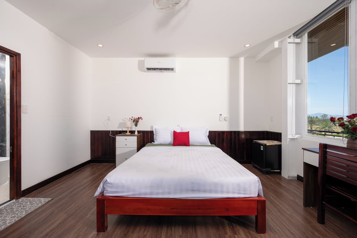 A modern bedroom with a large bed, a bedside table, a nightstand, a window with a view, and an air conditioning unit.