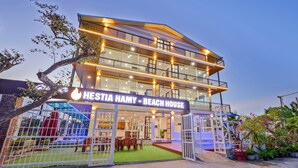 Een twee verdiepingen tellend strandhuis met een balkon en een overdekt terras, met een bordje waarop staat 'Hestia Hamy - Beach House'.