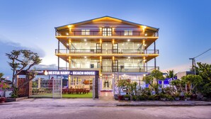 Een twee verdiepingen tellend strandhuis met een balkon, een overdekte zithoek buiten, en een bord met de tekst 'HESTIA HAMY - BEACH HOUSE'.