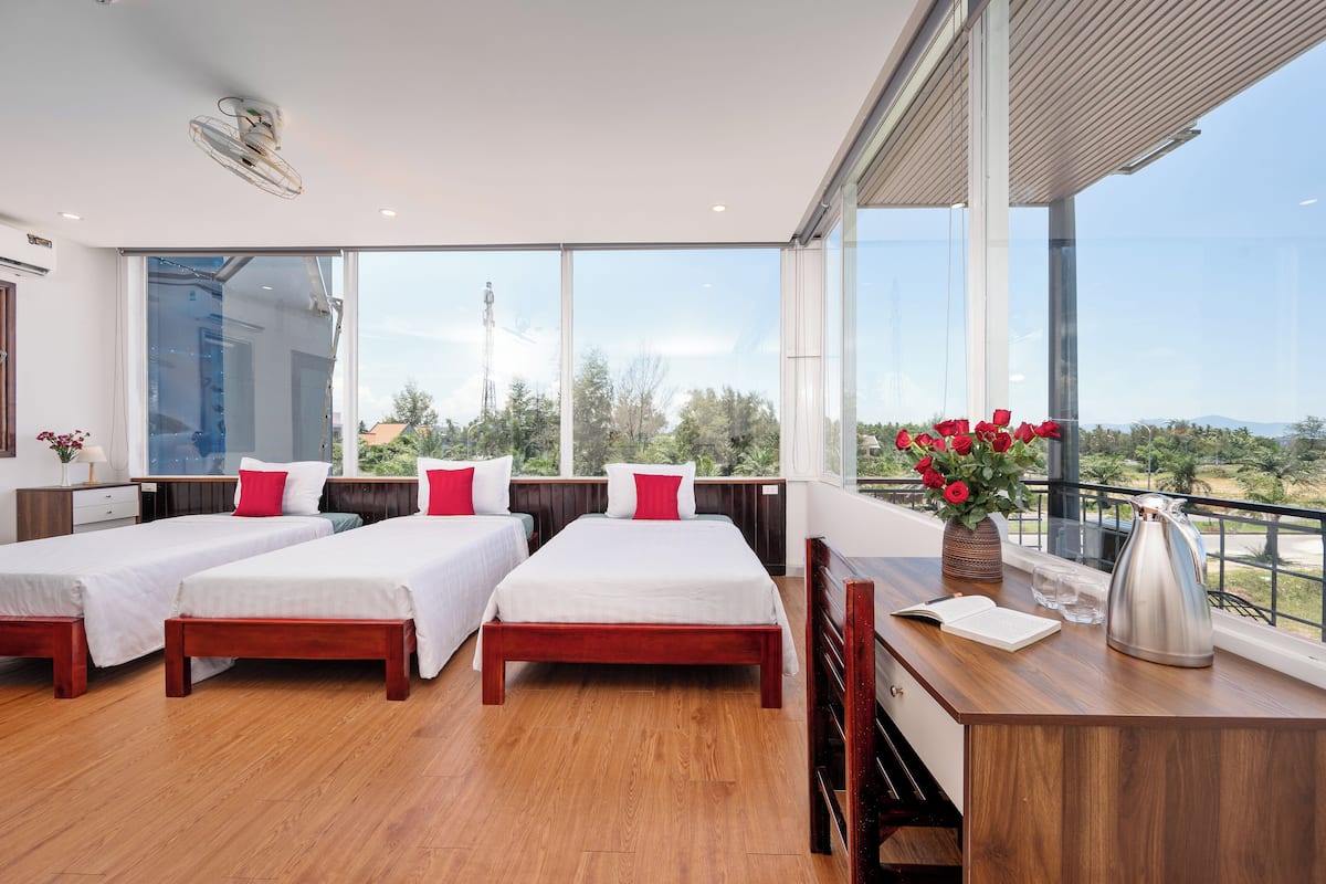 A modern hotel room with three beds, a wooden desk, a vase of red flowers, and a view of the cityscape.