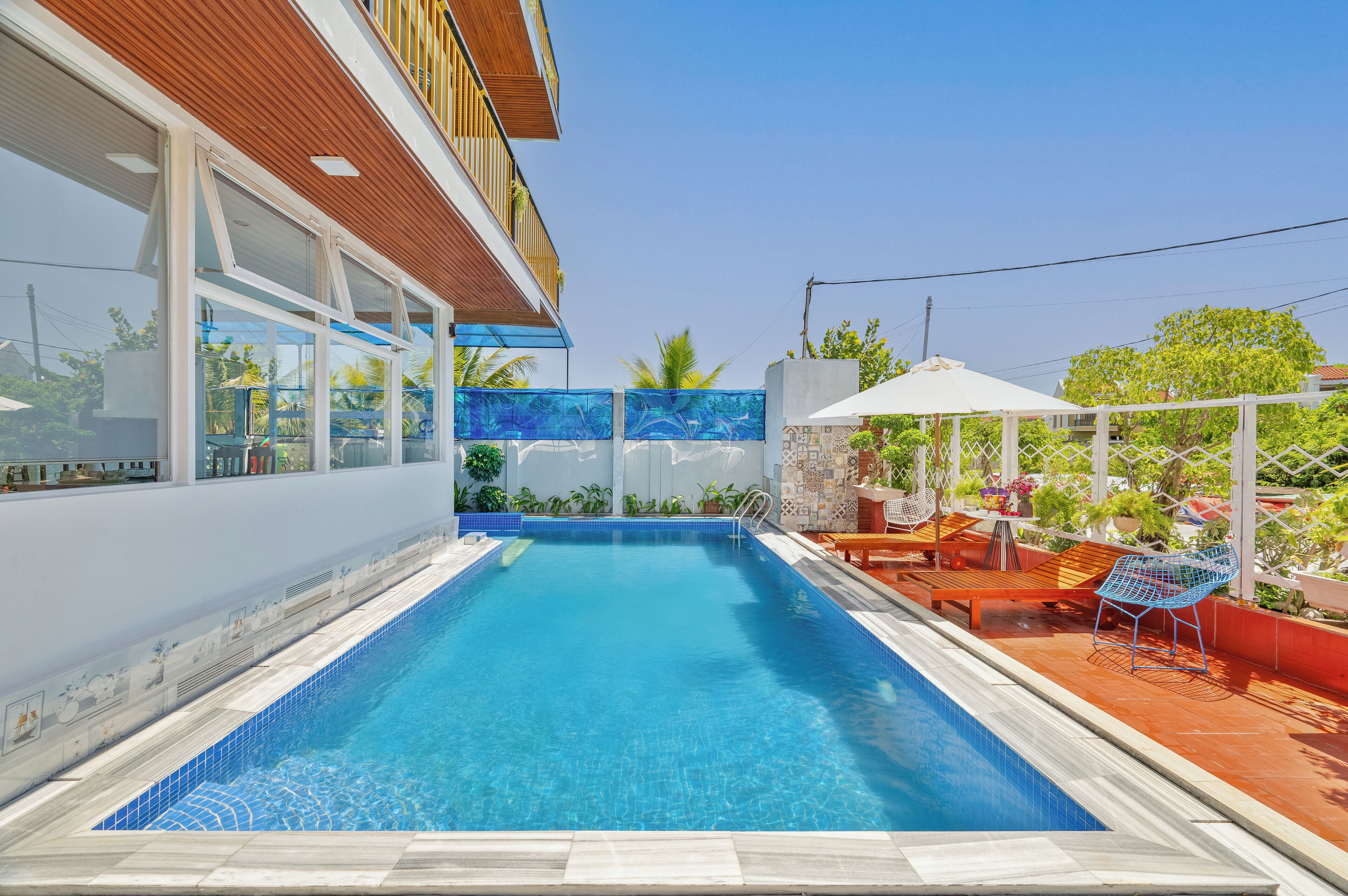 A swimming pool with a wooden deck and lounge chairs, surrounded by a white fence and plants.