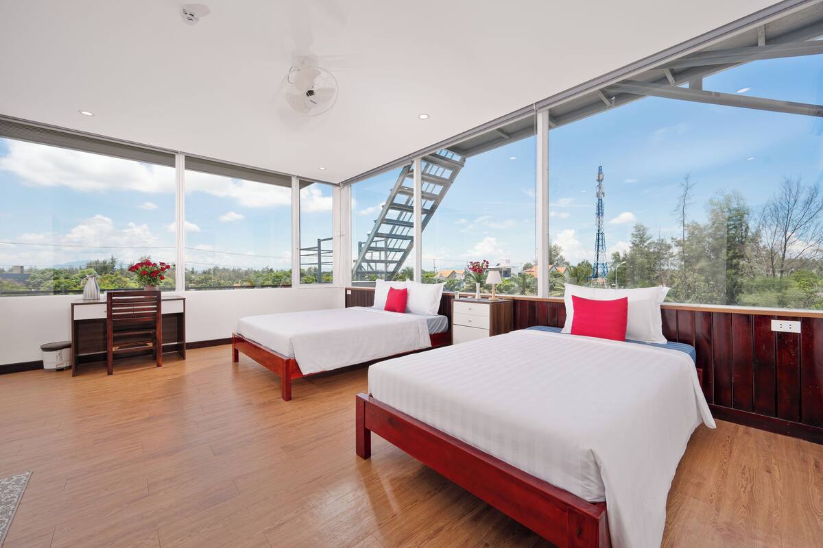 A modern hotel room with two beds, a desk, and large windows offering a view of the outdoors.