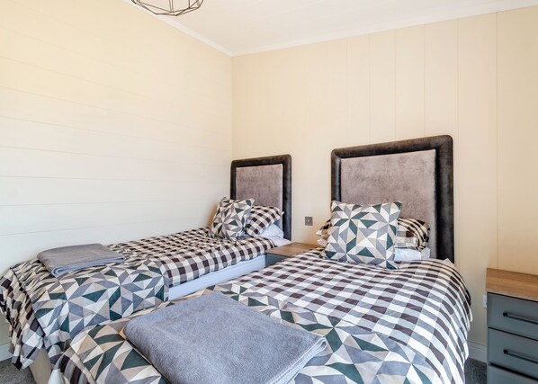 A room with two beds, each with a geometric patterned bedspread and matching pillows, a grey throw blanket, and a wooden nightstand.
