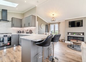 A modern kitchen with grey cabinets, a marble island, black bar stools, a built-in oven, and a TV above the fireplace.