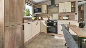 A modern kitchen with wooden cabinets, a stove, microwave, and a dining area with a table and chairs. - Platinum Caravan (Pet) (Bedale)