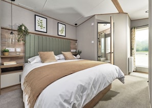 A modern bedroom with a bed, bedside tables, a green headboard, framed botanical prints, and a view of the bathroom.