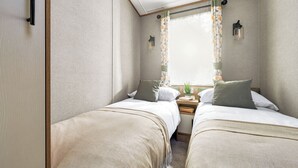A compact room with two beds, a small table, and a window with curtains. - Platinum Caravan (Pet) (Bedale)