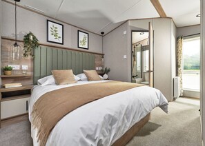 Ein modernes Schlafzimmer mit Bett, Nachttischen, einem grünem Kopfteil, gerahmten Pflanzenbildern und Blick ins Bad.
