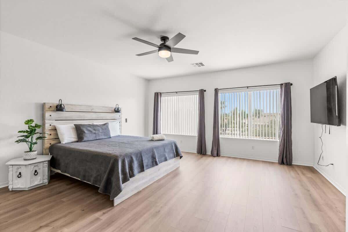 Une chambre à coucher comprenant un lit, une table de chevet, un ventilateur de plafond, un téléviseur et deux fenêtres avec des rideaux.