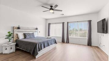 Une chambre à coucher comprenant un lit, une table de chevet, un ventilateur de plafond, un téléviseur et deux fenêtres avec des rideaux.