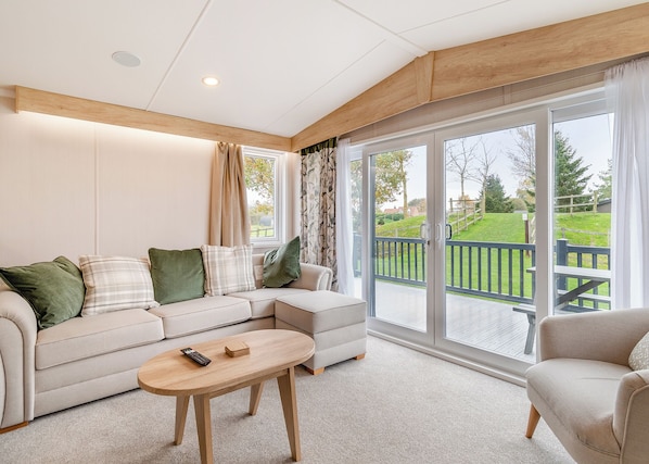 A modern living room with a sectional sofa, a wooden coffee table, and a view of a green landscape through large sliding glass doors. - Family and dog friendly lodge. (Swinethorpe, Nr Lincoln)