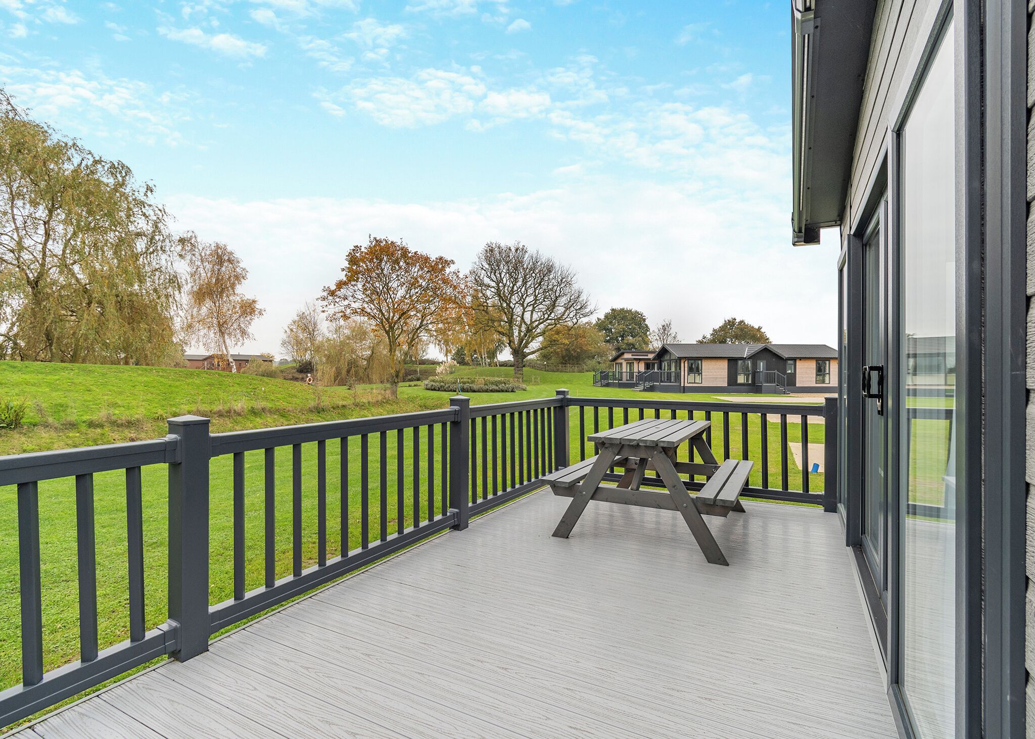 Une terrasse en bois avec une table de pique-nique, une rambarde noire et une vue sur un bâtiment et des arbres en arrière-plan.