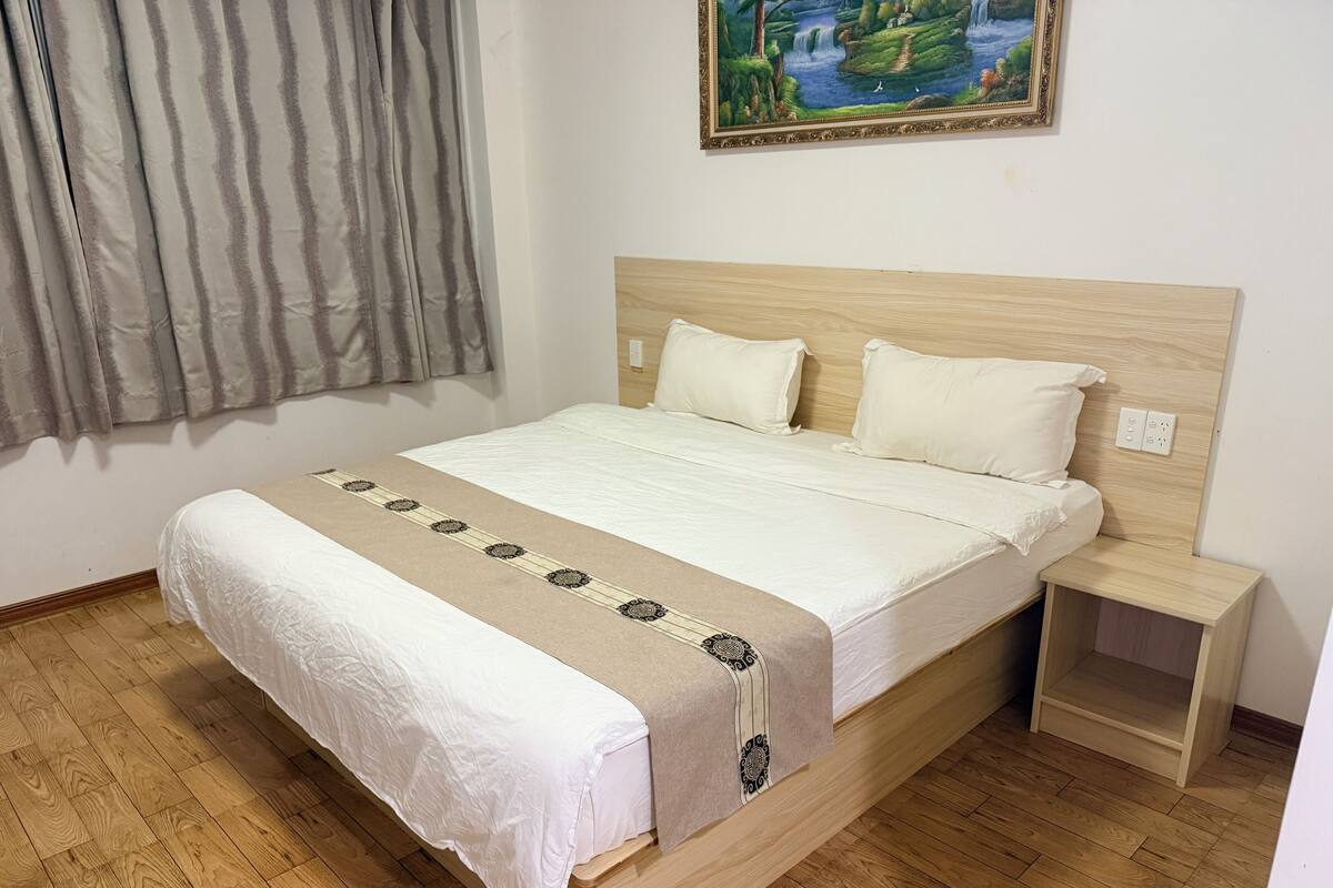 A neatly made bed with a patterned bedspread, a wooden headboard, a nightstand, and a painting of a landscape on the wall.