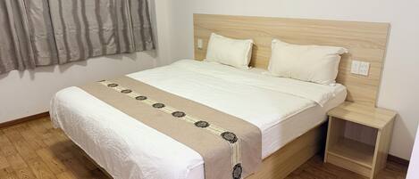 A neatly made bed with a patterned bedspread, a wooden headboard, a nightstand, and a painting of a landscape on the wall.