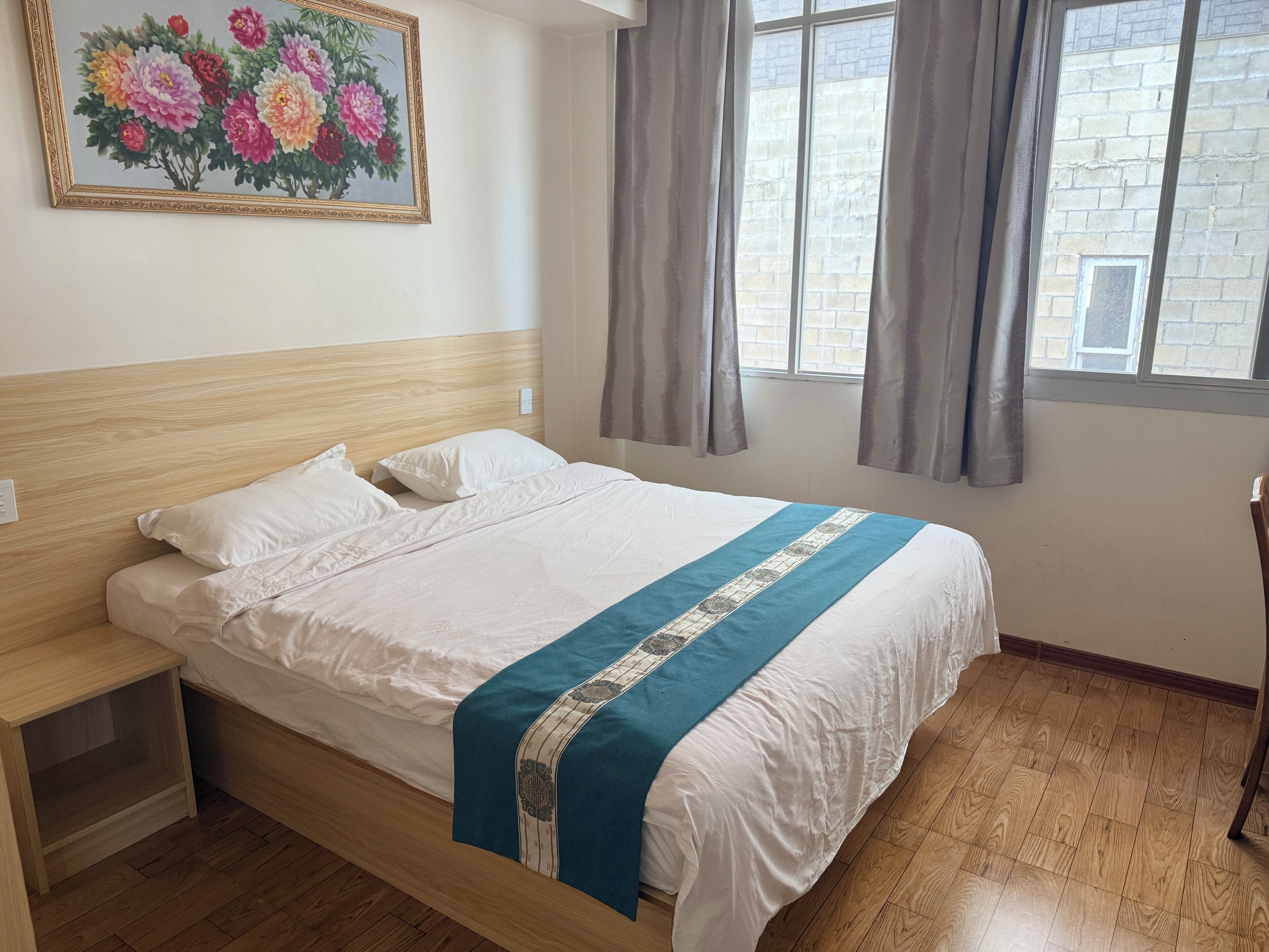 A neatly made bed with a blue and white bedspread, a wooden headboard, a nightstand, and a painting of flowers on the wall.
