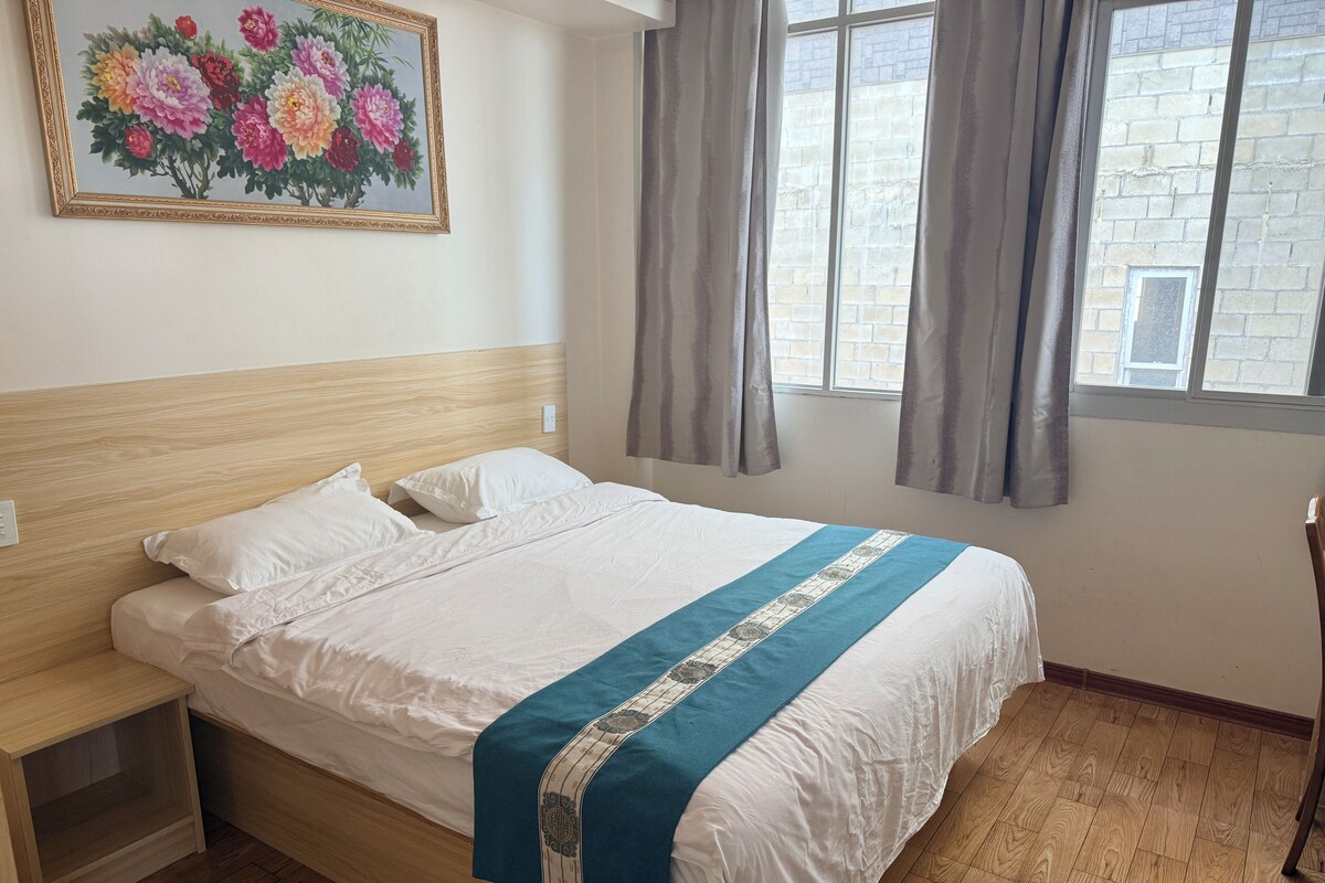 Ein ordentlich bezogenes Bett mit einer blau-weißen Tagesdecke, einem Holzkopfteil, einem Nachttisch und einem Blumengemälde an der Wand.