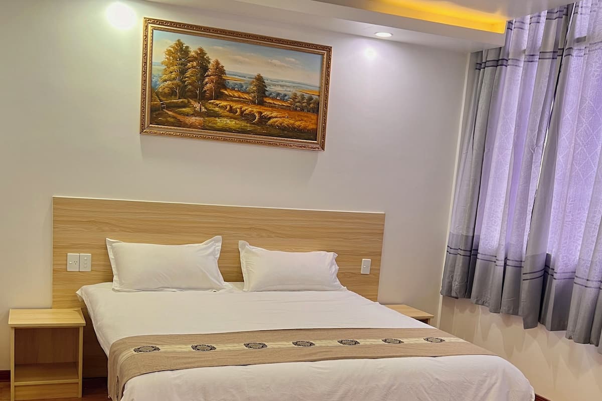 A bedroom with a wooden bed, white bedding, a nightstand, a painting on the wall, and a window with curtains.