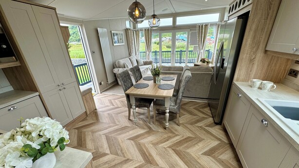 A modern kitchen with a herringbone floor, a dining area with a wooden table and grey upholstered chairs, and a view of a balcony with greenery. - Platinum 2 Bed Spa Slps 4 (Pet) (Clarach)