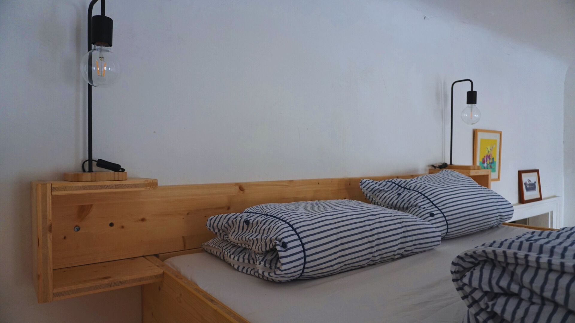 Un lit simple avec une literie à rayures, une tête de lit en bois, une table de chevet avec une lampe et un luminaire fixé au mur.