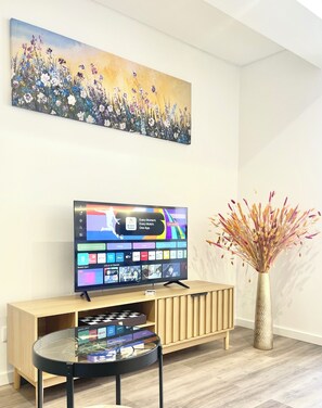 A living room with a flat-screen TV on a wooden stand, a glass coffee table, a decorative vase with dried plants, and a painting of flowers on the wall. - Luxury T3 Plaza Inn | A/C, Lift, Balcony in Town Centre (Funchal)