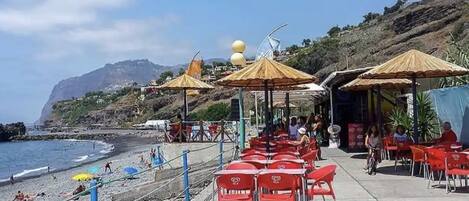 Un café frente a la playa con terraza y sombrillas, con vistas al océano y a un paisaje montañoso.