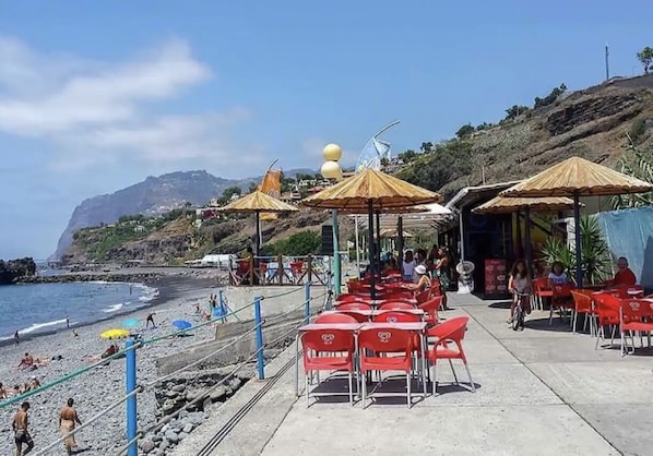 Un café frente a la playa con terraza y sombrillas, con vistas al océano y a un paisaje montañoso.