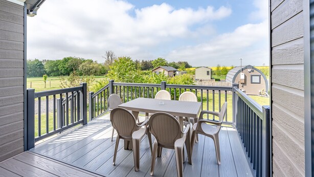 A covered outdoor deck with a table and chairs, a view of a green landscape, and a wooden building in the background. - Aura Chalet (Pet Friendly) (Coldstream)