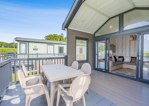 A modern outdoor deck with a table and chairs, a sliding glass door leading to an interior seating area, and a view of a green mobile home in the background. - Aura Chalet (Pet Friendly) (Coldstream)