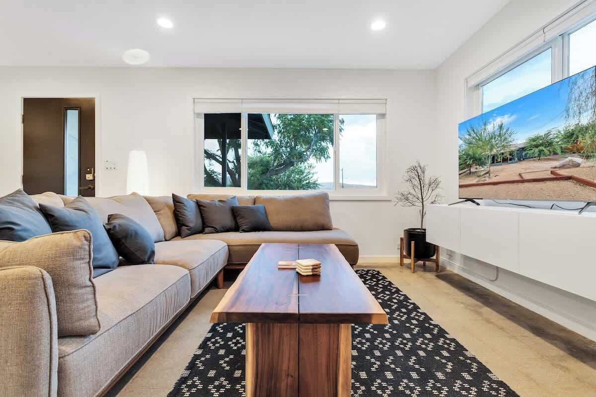 Een moderne woonkamer met een hoekbank, een houten salontafel en een groot raam met uitzicht op bomen en een gebouw.