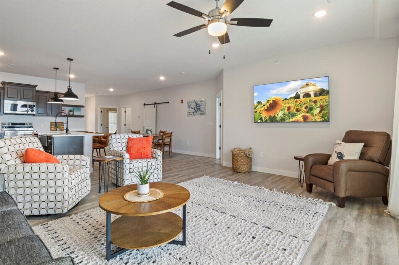 A modern living room with a sofa, armchair, and coffee table, a dining area with chairs, a kitchen with appliances, and a ceiling fan.