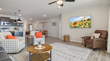 A modern living room with a sofa, armchair, and coffee table, a dining area with chairs, a kitchen with appliances, and a ceiling fan.