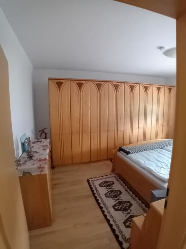 A bedroom with a wooden wardrobe, a bed with a patterned bedspread, and a small table with a floral tablecloth.