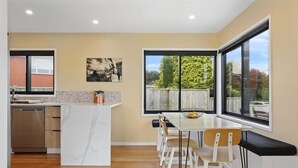 A modern kitchen with a marble island, wooden cabinets, and a dishwasher. There is a dining area with a table and chairs, and large windows offering a view of the outdoors. - Sundrenched 70's - Spa Pool + Sleep-out, Sleeps 11 (Rotorua)