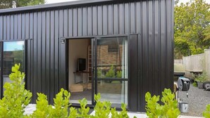 A modern building with a dark exterior, large glass sliding doors, and a visible interior with a ladder and a table. - Sundrenched 70's - Spa Pool + Sleep-out, Sleeps 11 (Rotorua)