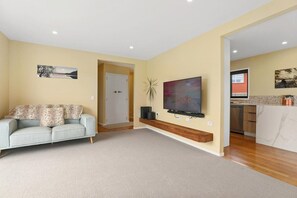 A living room with a sofa, a flat-screen TV, and a kitchen area in the background.