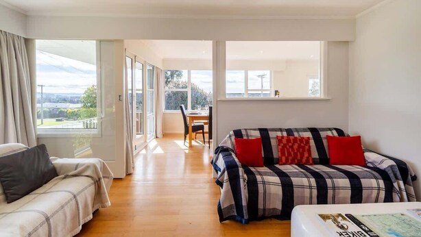 A modern room with a sofa, a dining table, and a view through large windows. - All Seasons Escape - Hilltop, Sleeps 10, Bikes (Rotorua)