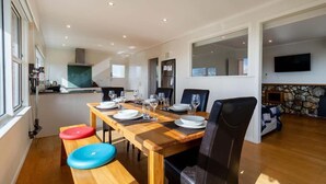 A dining area with a wooden table set for four, a kitchen in the background, and a stone wall with a television. - All Seasons Escape - Hilltop, Sleeps 10, Bikes (Rotorua)