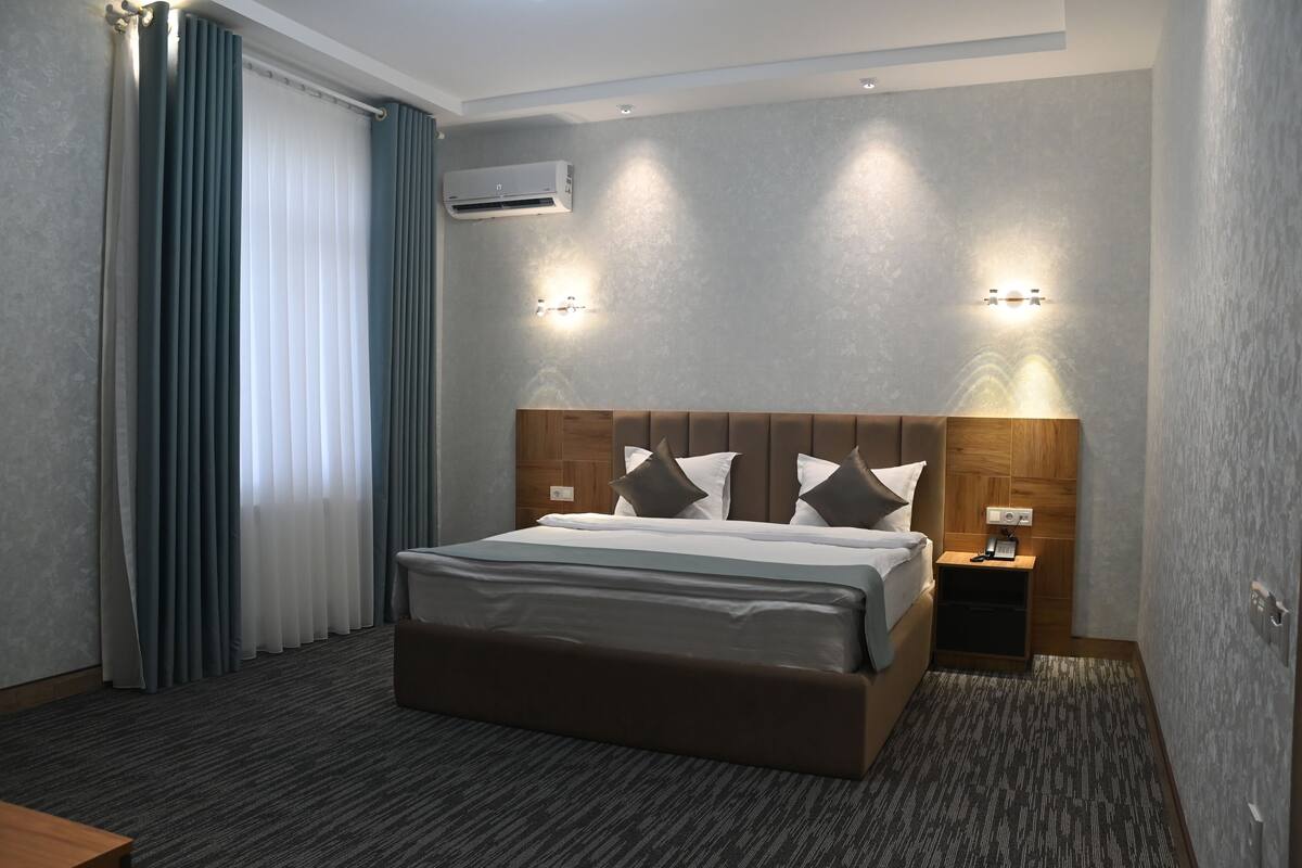 A modern hotel room with a large bed, a nightstand, an air conditioning unit, and a wall-mounted light fixture.